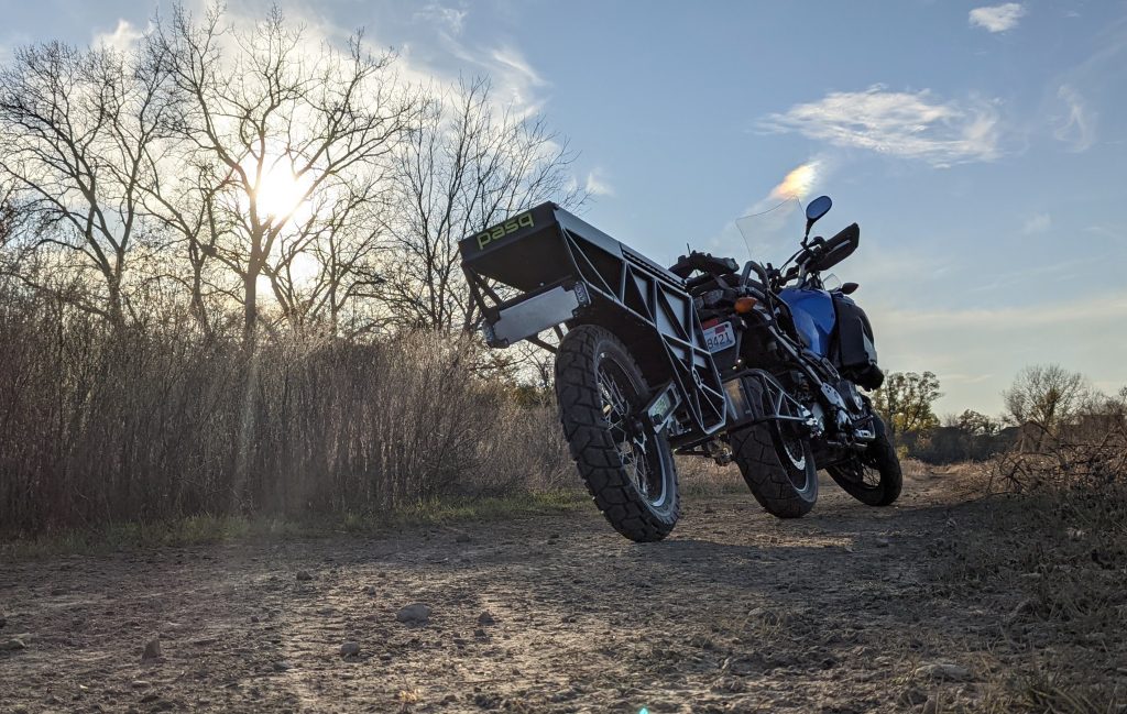 The Pasq ADV1, a single-wheeled dual-sport and Adventure trailer for motorcycles, on a dirt road with the sun setting in the background.