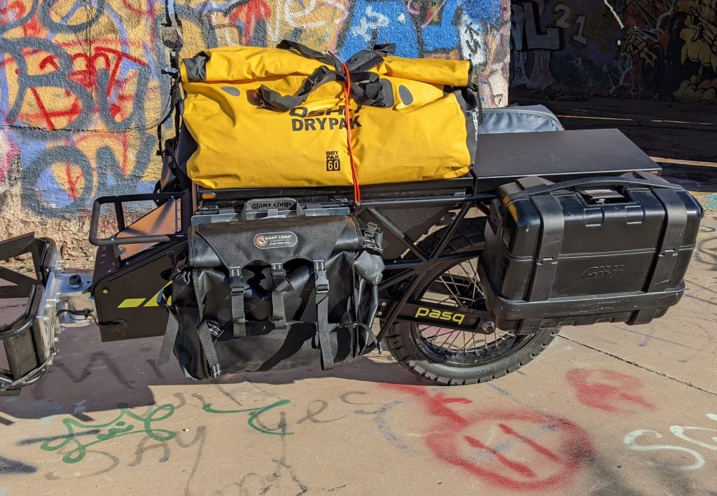 The Pasq ADV1 dual-sport adventure motorcycle trailer decked out with a yellow dry bag, a GiantLoop Round The World Pannier, a Givi Mono-Key pannier, all against a graffitied building background.